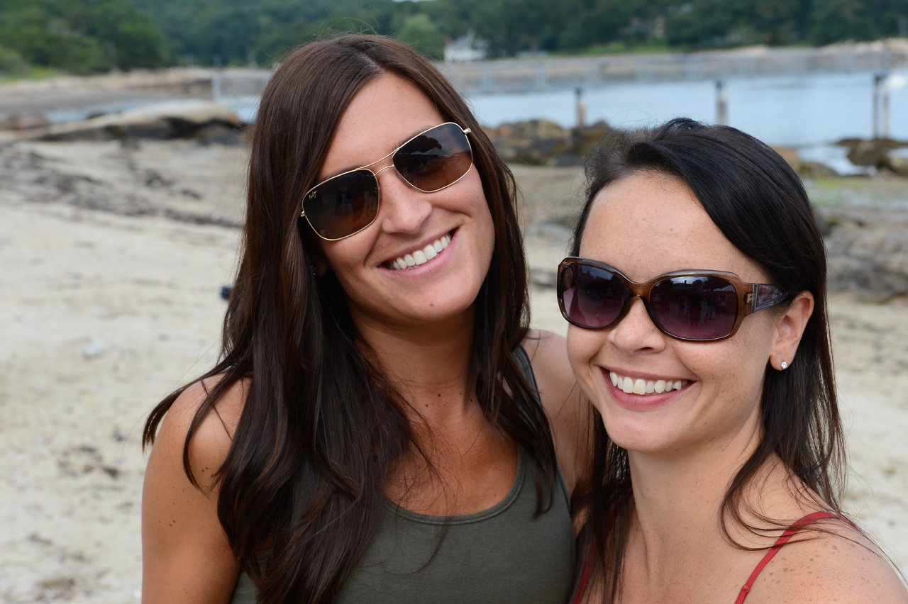 Two smiling women wearing sunglasses pose together on a sandy beach with water and rocks in the background.