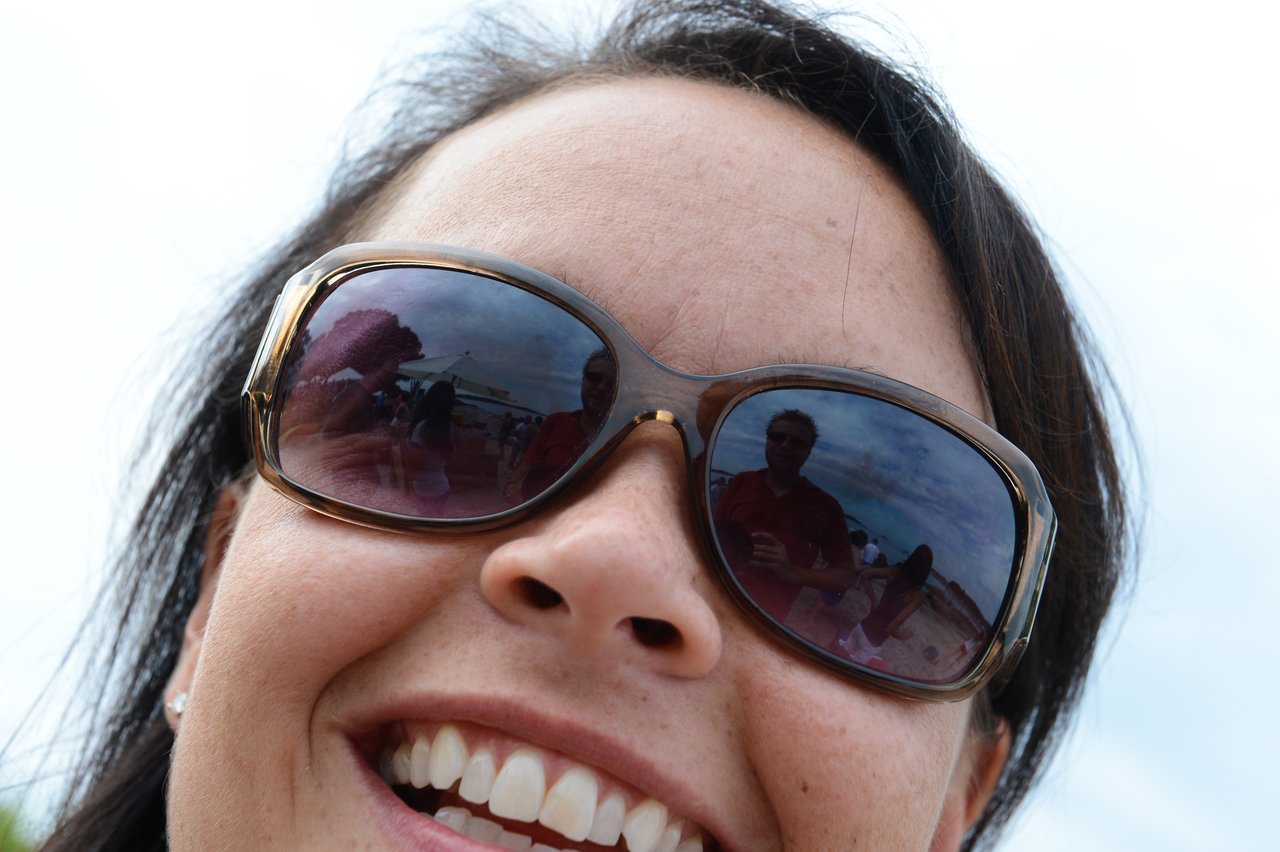 Close-up of a smiling person wearing sunglasses, reflecting a group of people at an outdoor party.