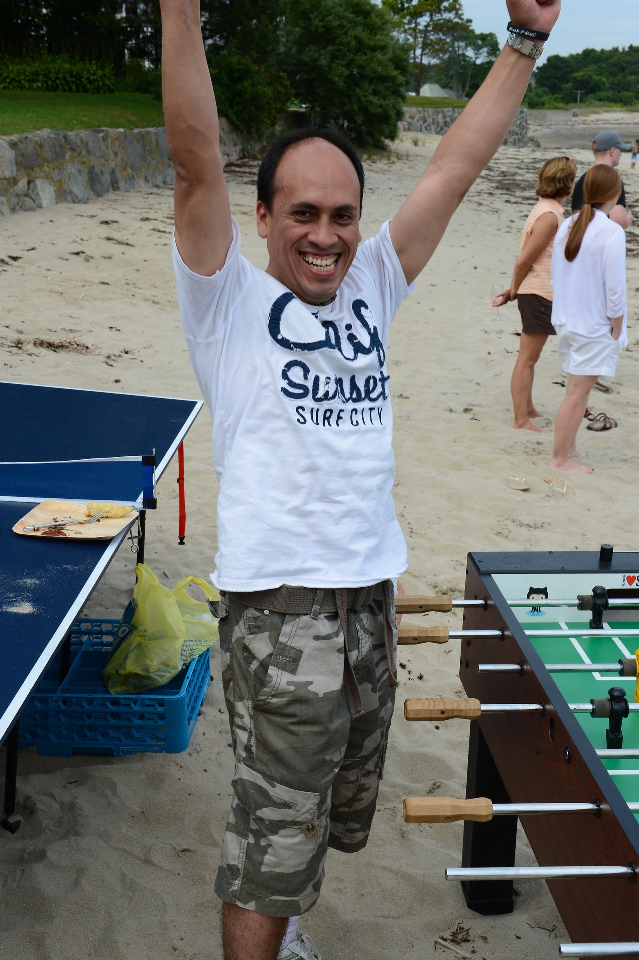 A man on a sandy beach raises his arms and smiles near a foosball table and ping pong table.