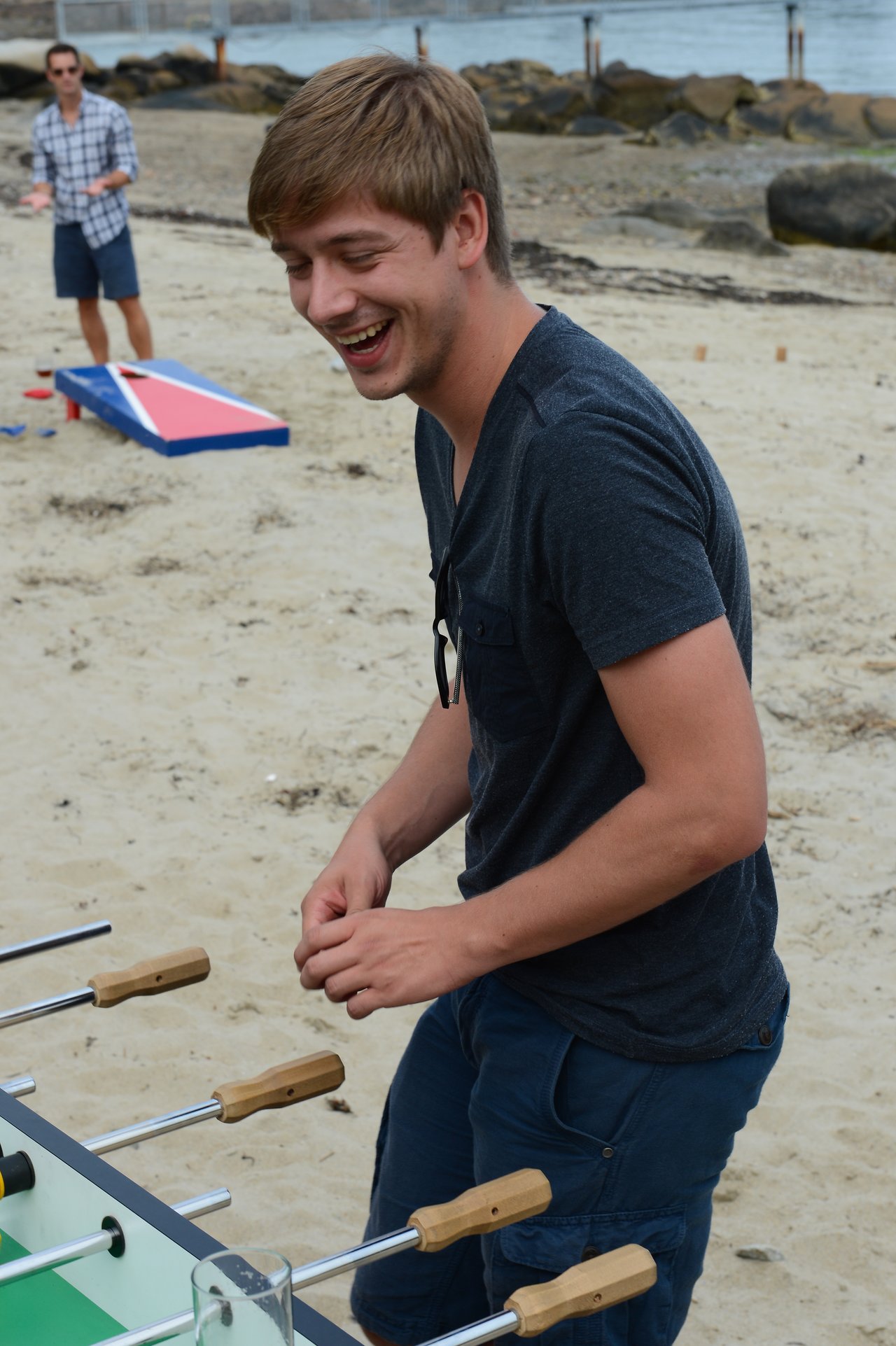 A man laughs while playing foosball on a sandy beach, with another person playing cornhole in the background.