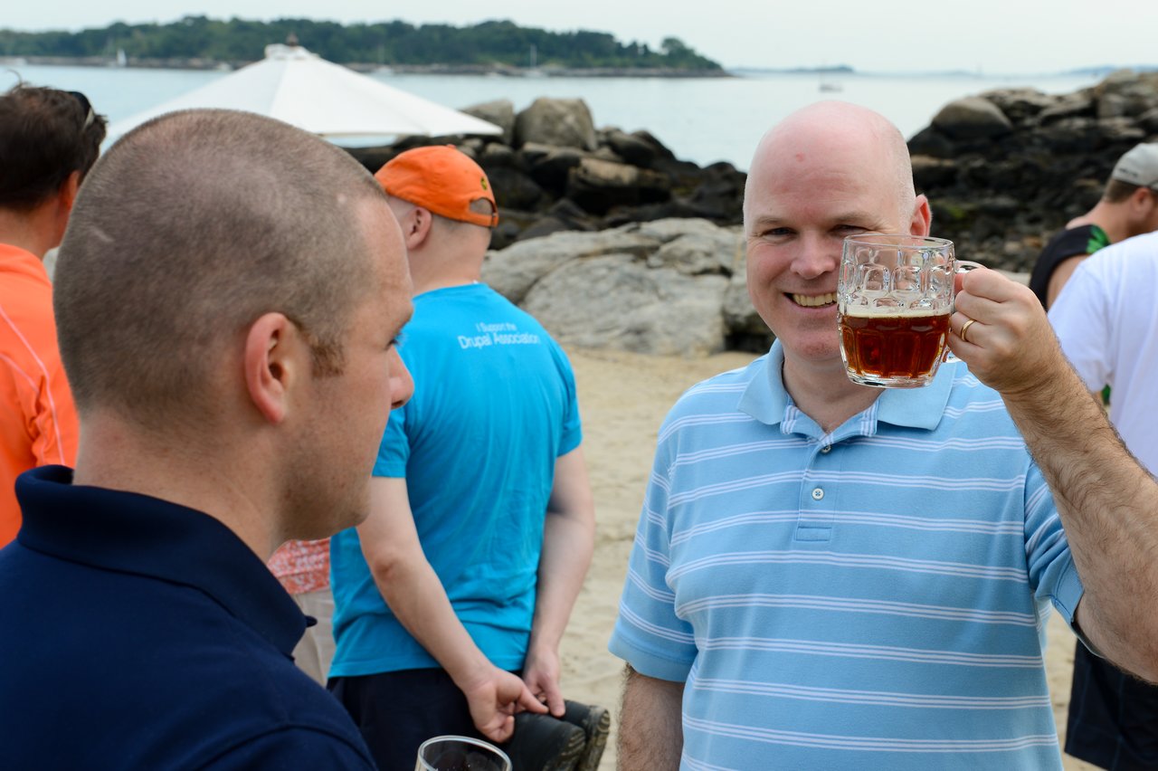 A smiling man in a blue striped shirt holds up a beer mug at an outdoor beach gathering.