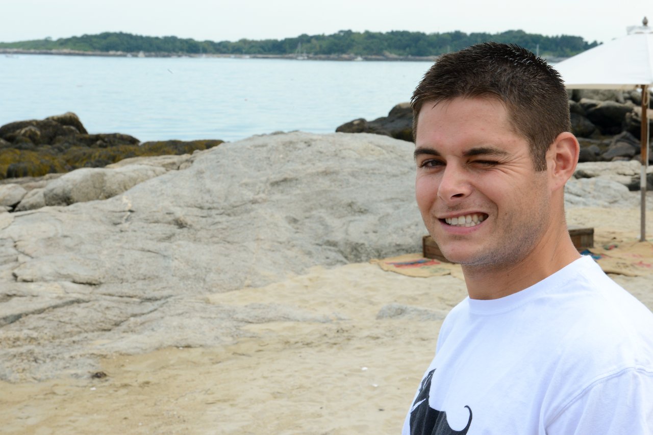 A man in a white t-shirt winks and smiles while standing on a sandy beach with rocks and water.