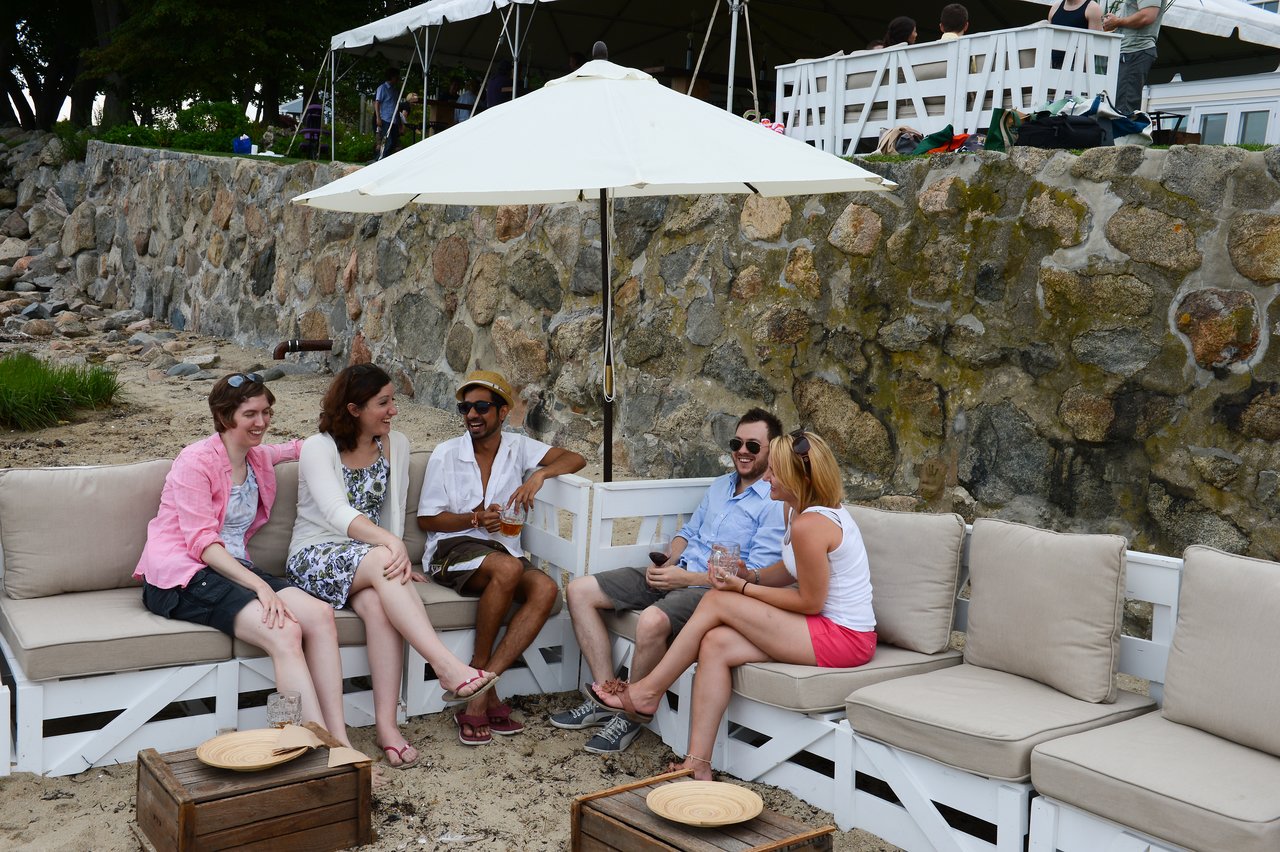 A group of people sit on outdoor couches, chatting and laughing at a summer party by the beach.