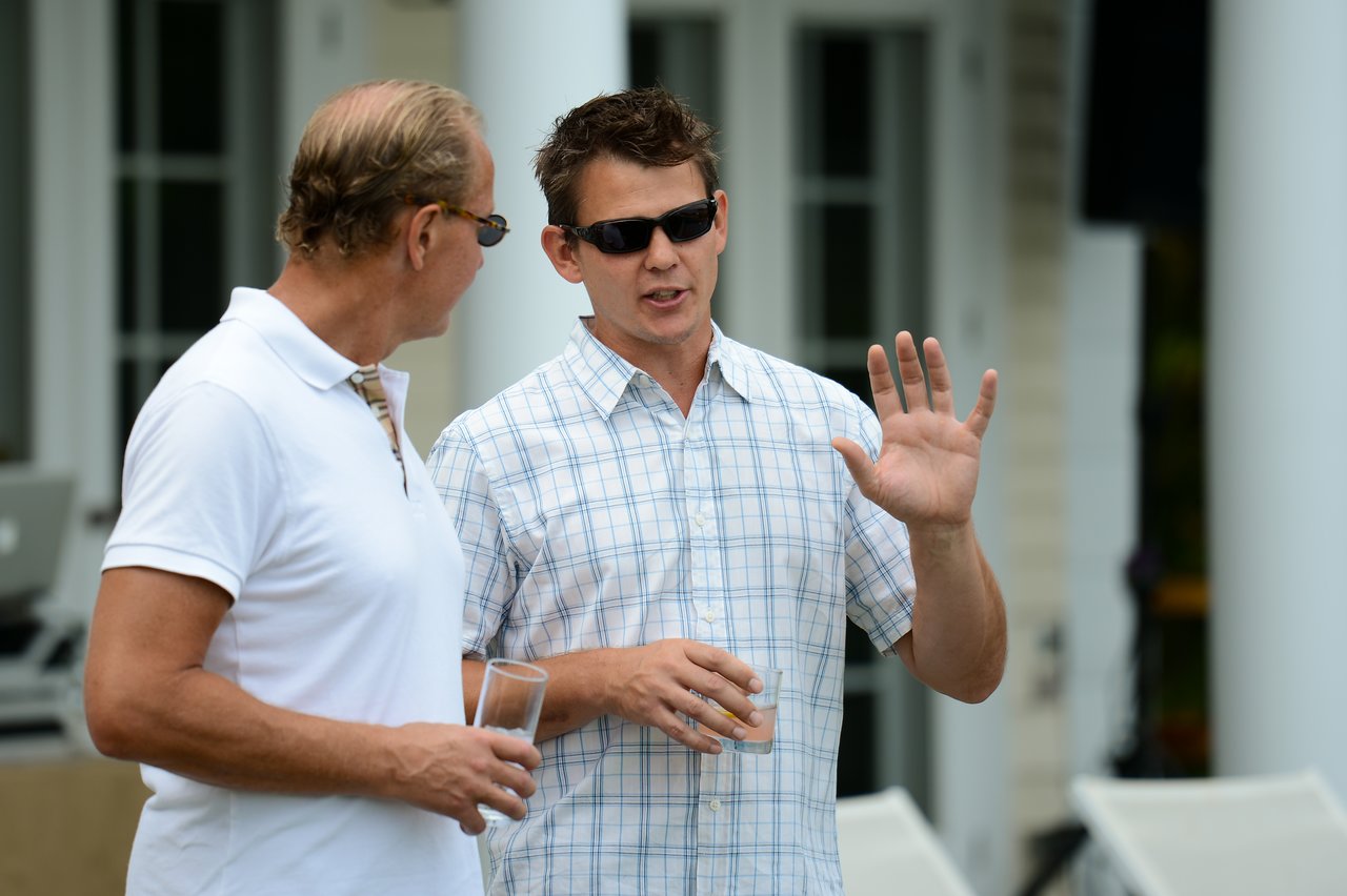 Two men wearing sunglasses talk outside, one gesturing with his hand while the other holds a drink.