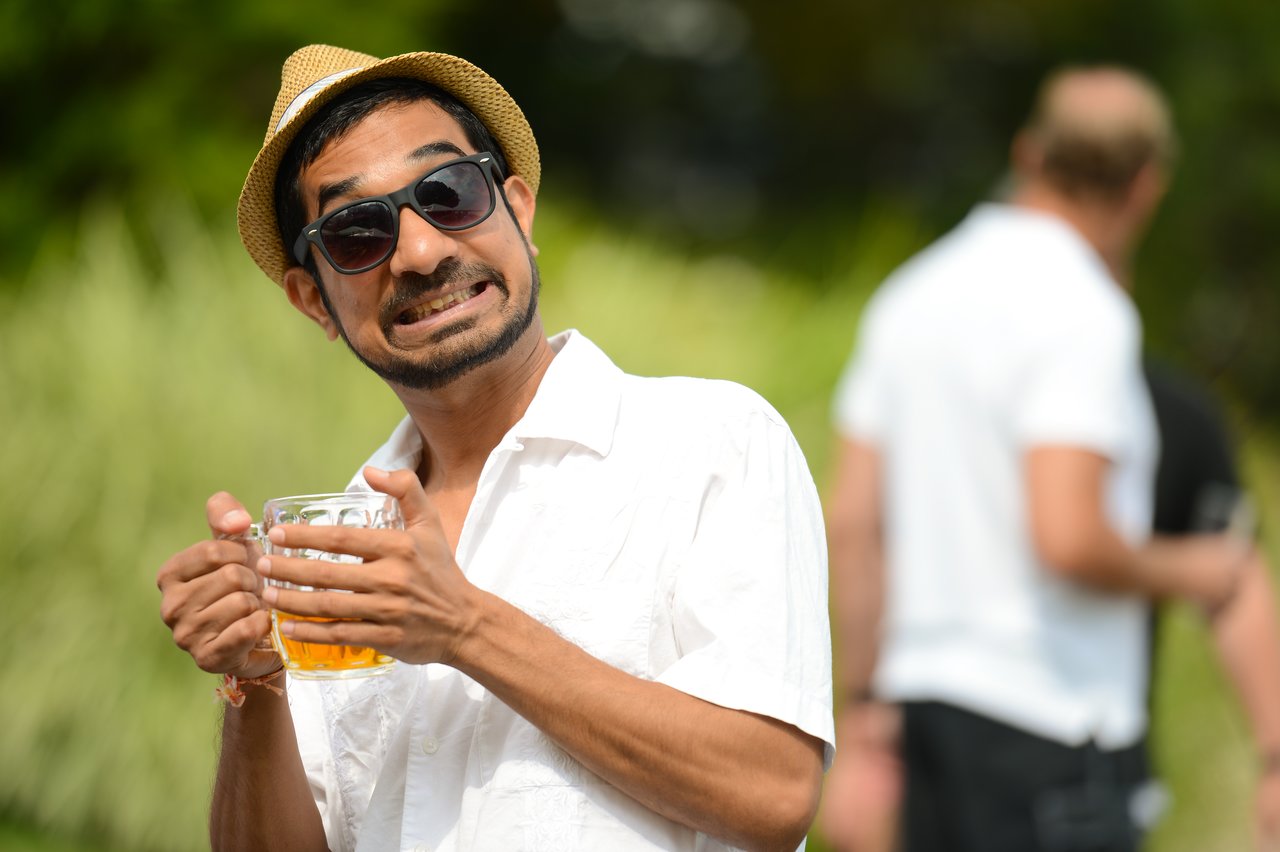 A man in a white shirt and hat smiles playfully while holding a glass of beer at an outdoor party.