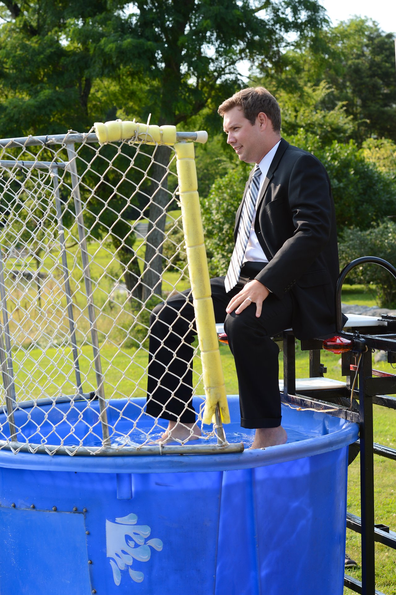 A man in a suit sits on a dunk tank platform, preparing to be dropped into the water.