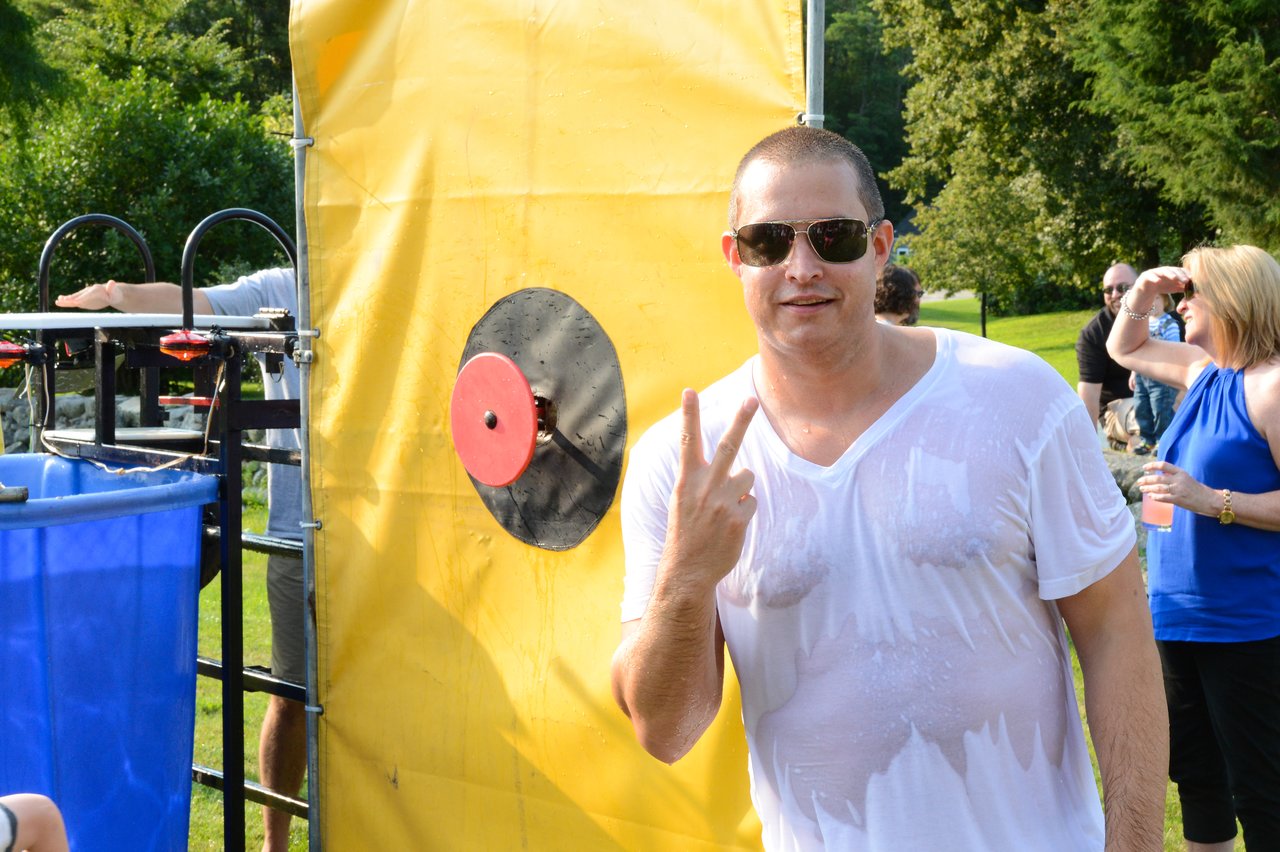 A man in a wet shirt and sunglasses stands near a dunk tank, holding up a peace sign.