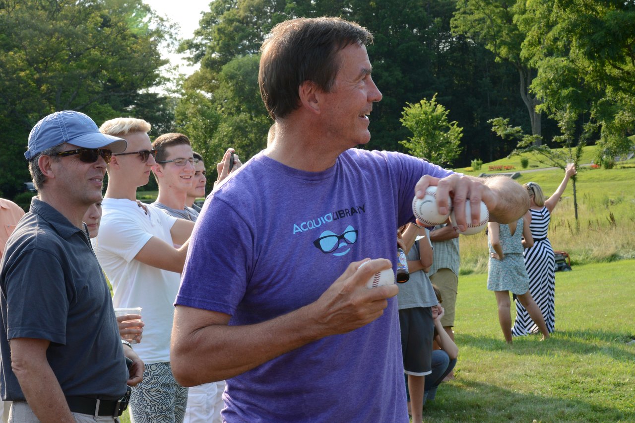 A man in a purple shirt holds baseballs, preparing to throw at a dunk tank target during an outdoor event.