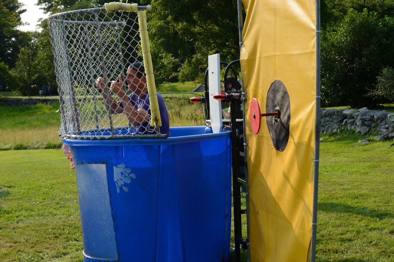 A person sits inside a dunk tank, gripping the cage, as the target is hit during an outdoor event.