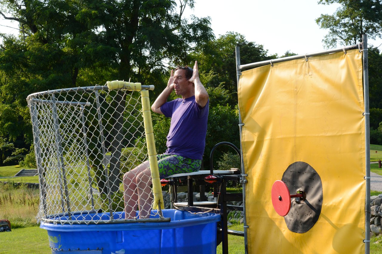 A person sits on a dunk tank platform, hands on head, waiting to be dropped into the water.