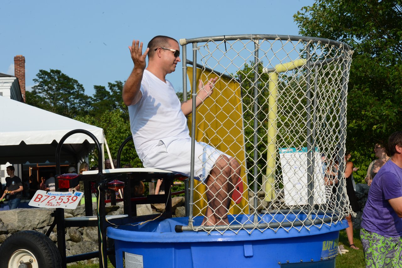 A man sits on a dunk tank seat, raising his hands as someone prepares to throw a ball.