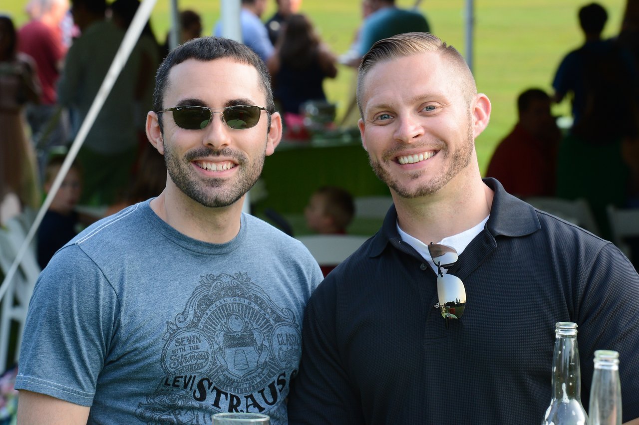 Two men smiling at the camera during an outdoor summer party, with drinks on the table in front of them.