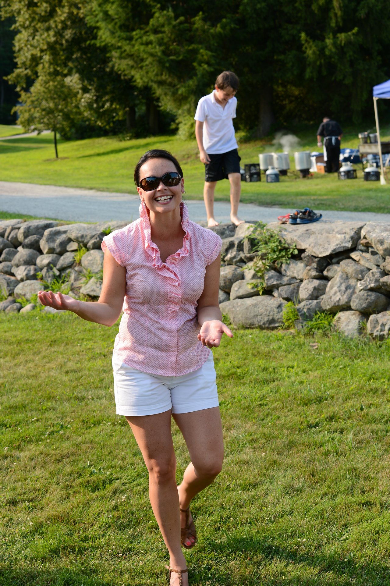 A smiling woman in sunglasses walks on the grass, gesturing with her hands at an outdoor summer gathering.