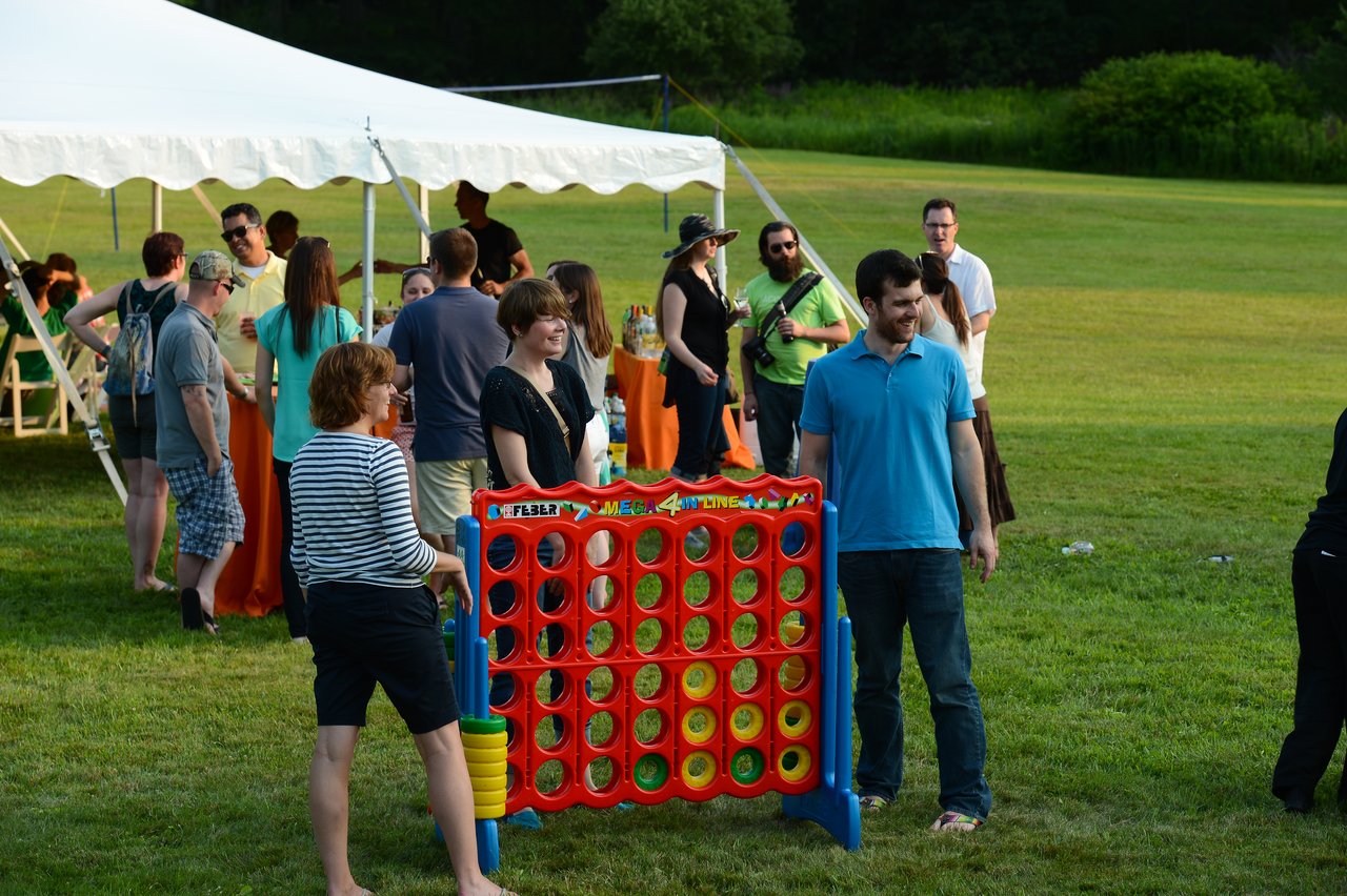People gathered outdoors at a summer party, playing a large Connect Four game and socializing near a tent.