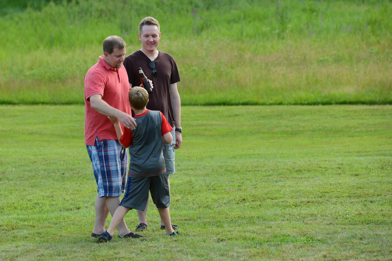 A man in a red shirt interacts with a young boy while another man watches, holding a drink.