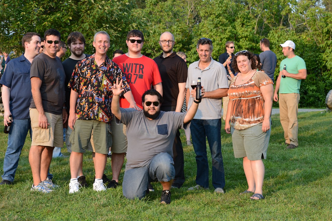 A group of people poses outdoors, smiling, with one person kneeling in front raising their hands and holding a drink.