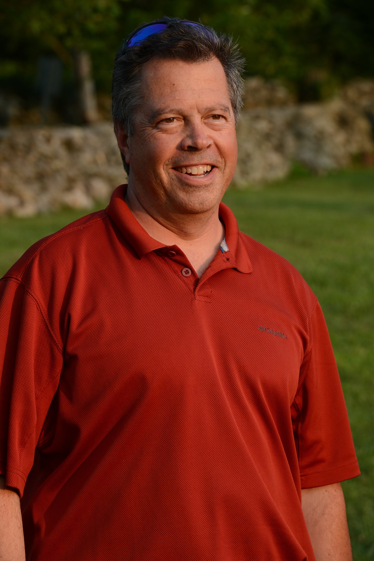 A man in a red polo shirt smiles while standing outdoors at a summer party.