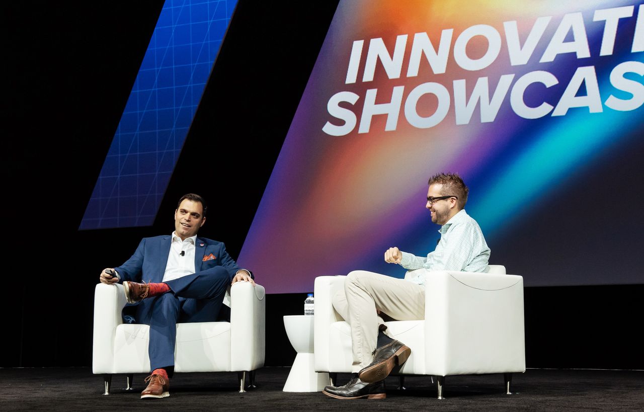 Two men sit on stage in white chairs, having a conversation during an Acquia Engage event interview.