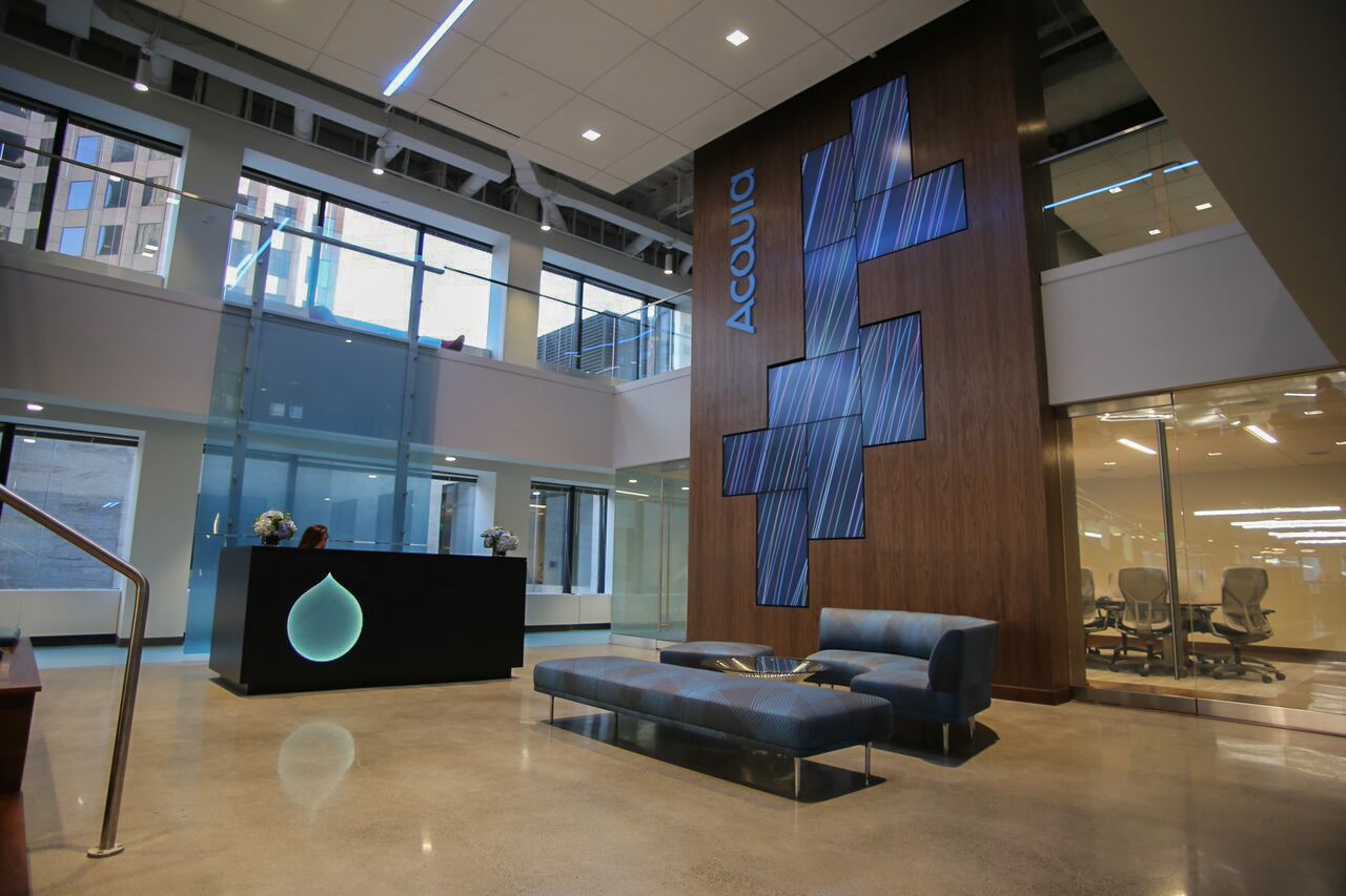A modern office lobby with a reception desk, seating area, and a large digital display on a wooden wall.