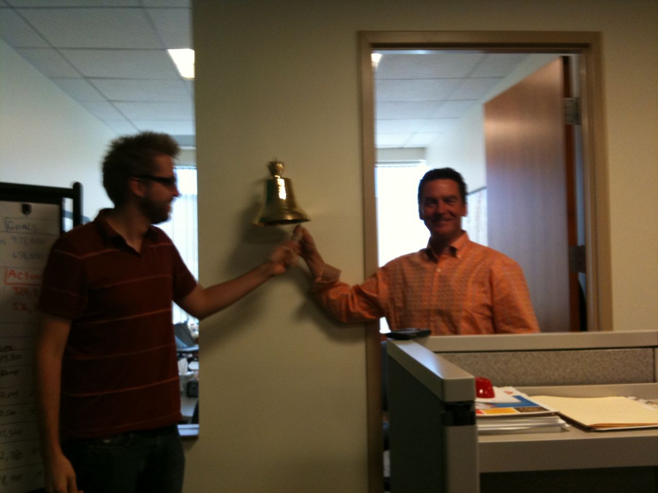 Two men smiling while ringing a bell mounted on a wall in an office.
