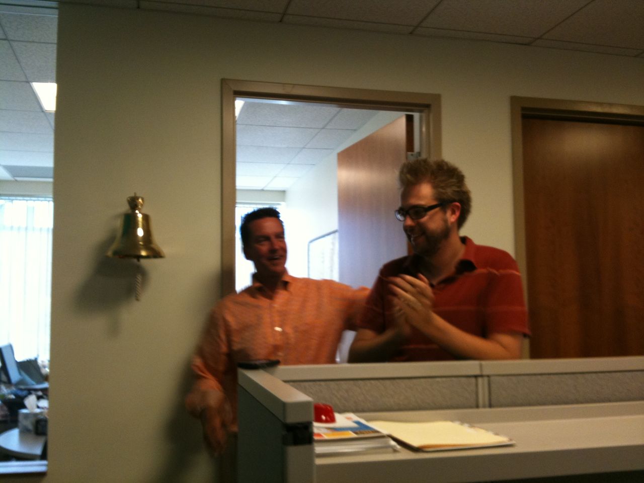 One man claps while the other smiles and gestures near a wall-mounted bell in an office setting.