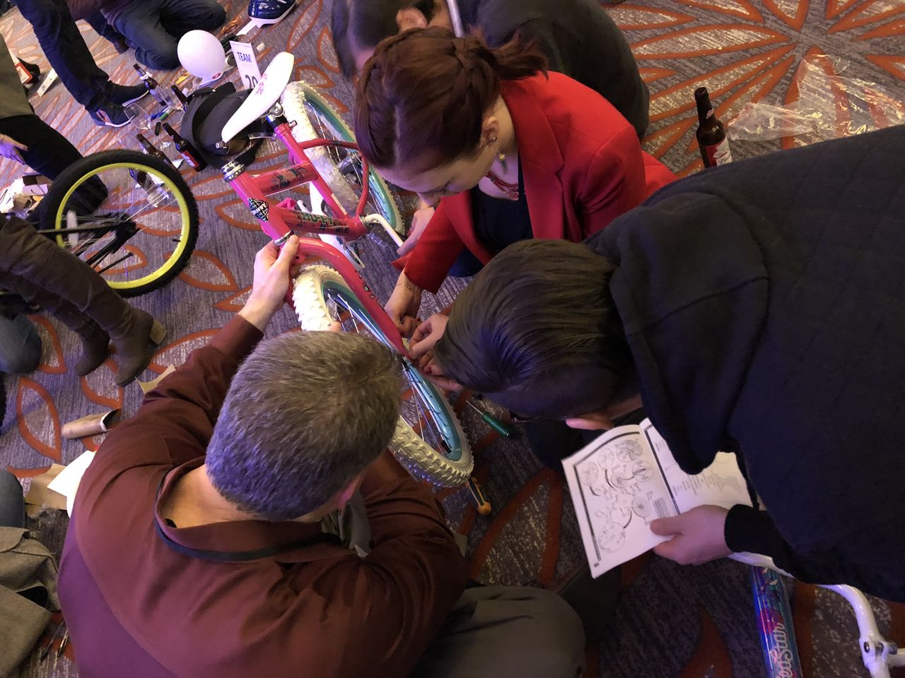 A group of people work together to assemble a pink bicycle, following instructions from a manual.