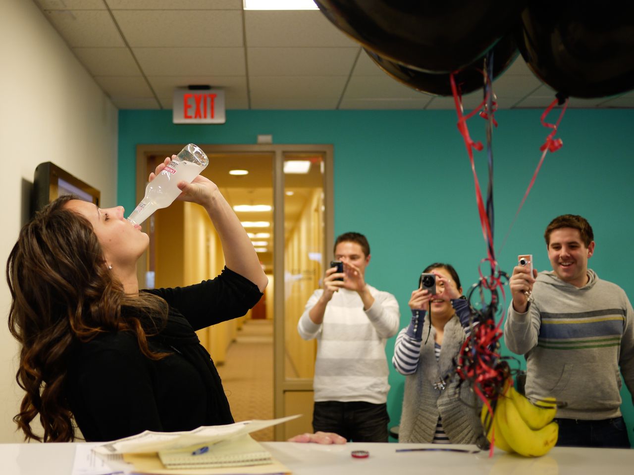 A woman drinks from a bottle while three people take photos and smile in a decorated office.