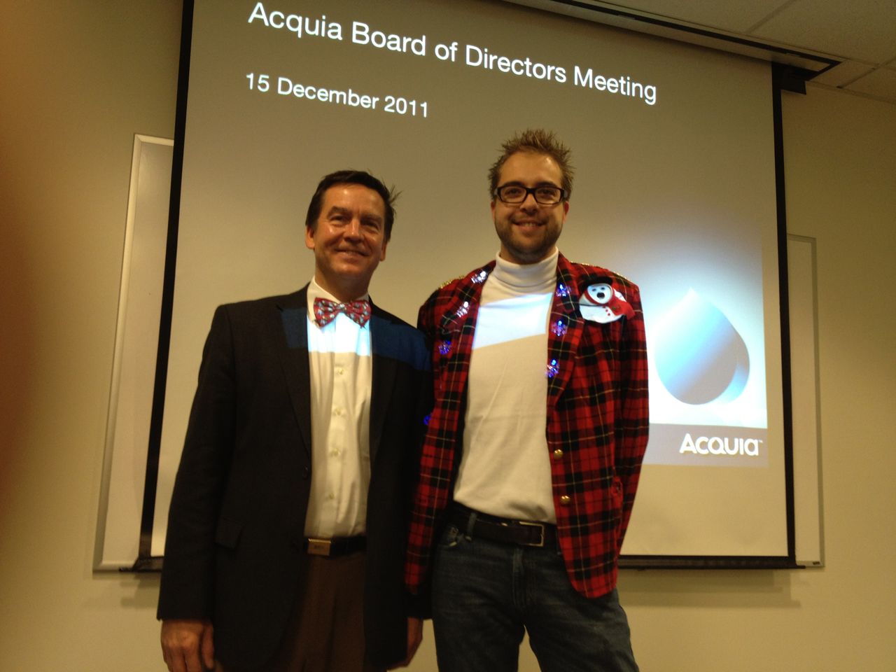 Two people smiling in festive attire at an Acquia board meeting, standing in front of a presentation screen.