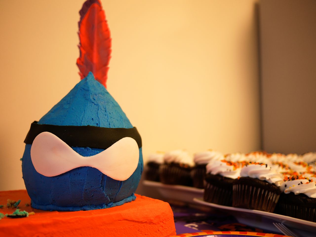 A blue, masked cake with a red feather sits on a table, with decorated cupcakes in the background.