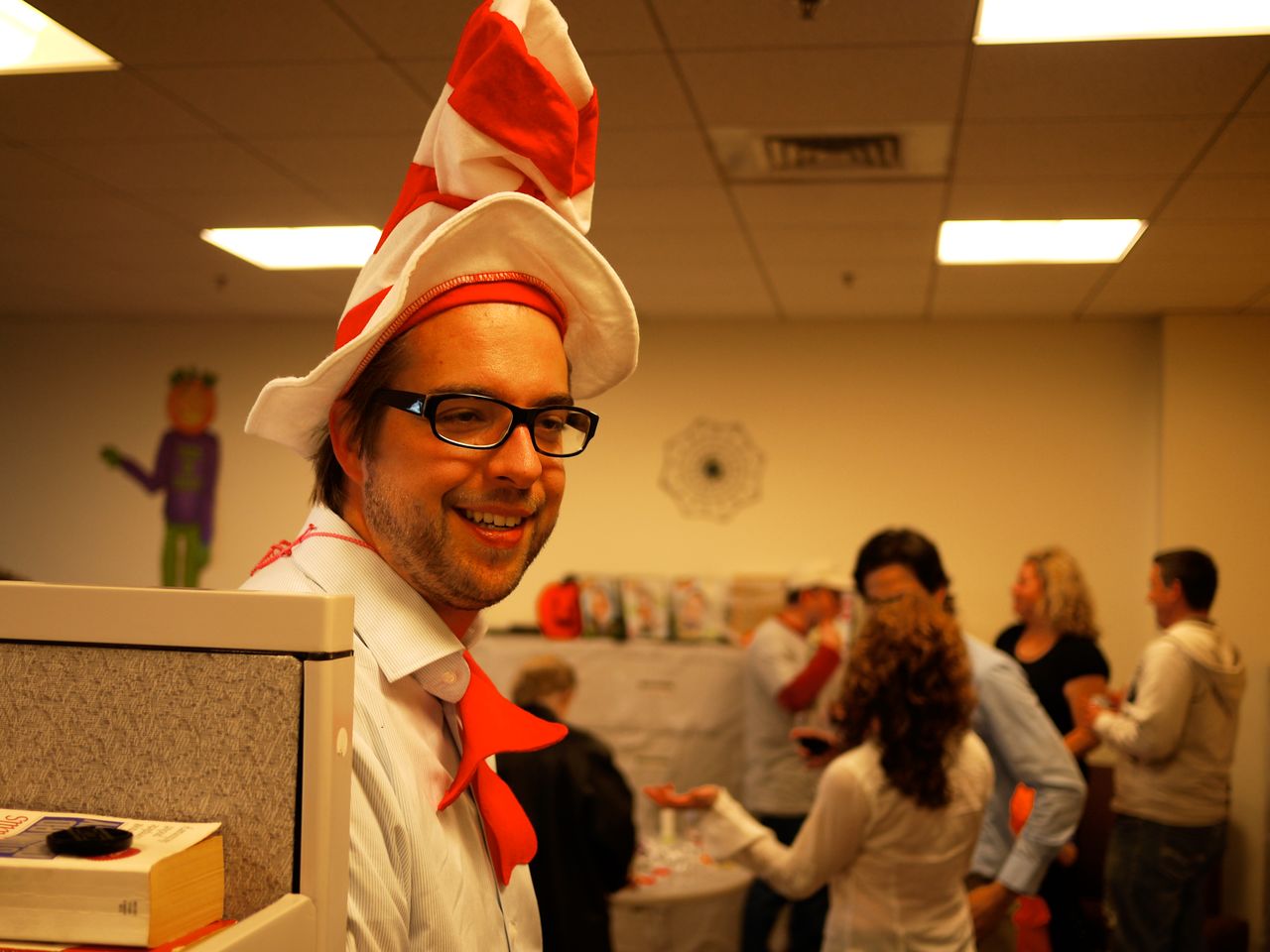 A man in a red and white costume hat smiles at the camera during a Halloween office gathering.