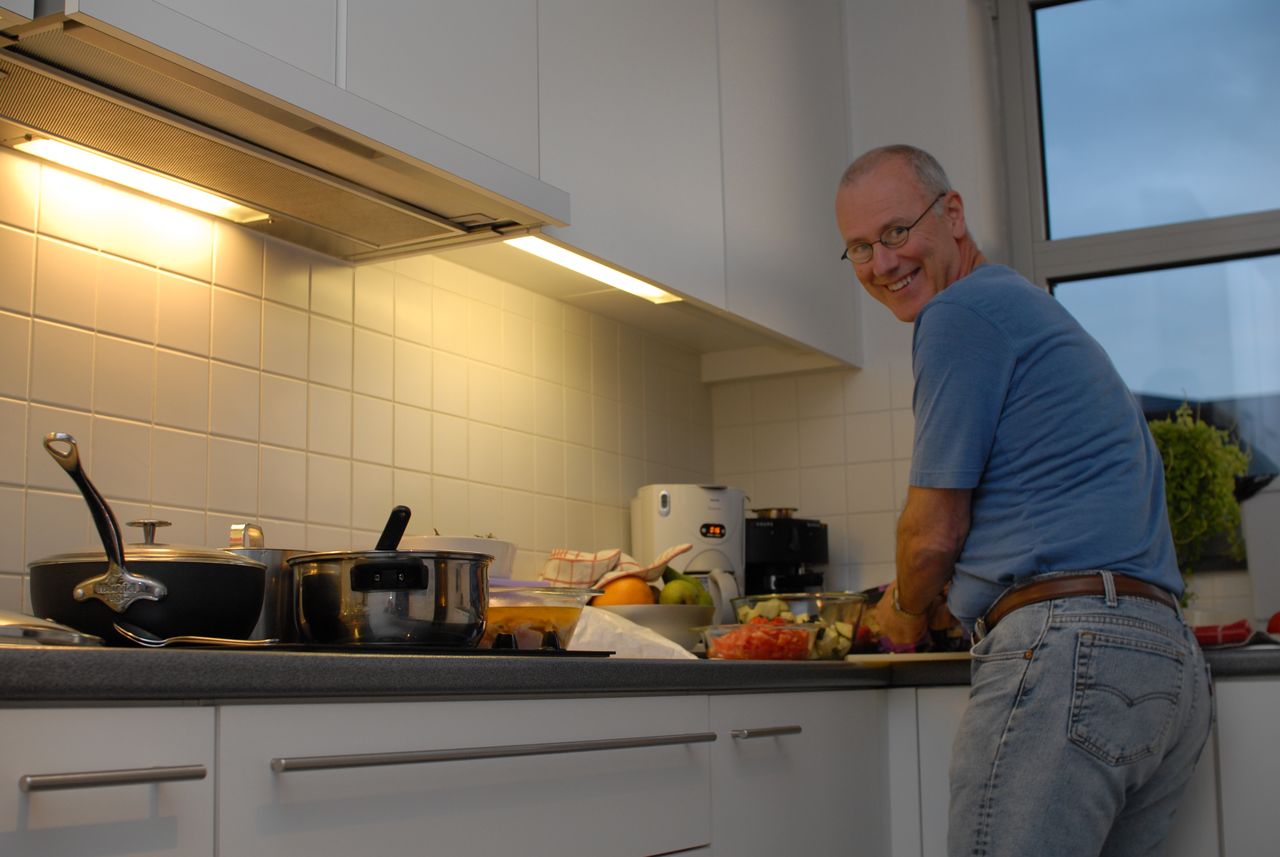 A man in a blue shirt smiles while preparing food in a kitchen with pots and ingredients on the counter.