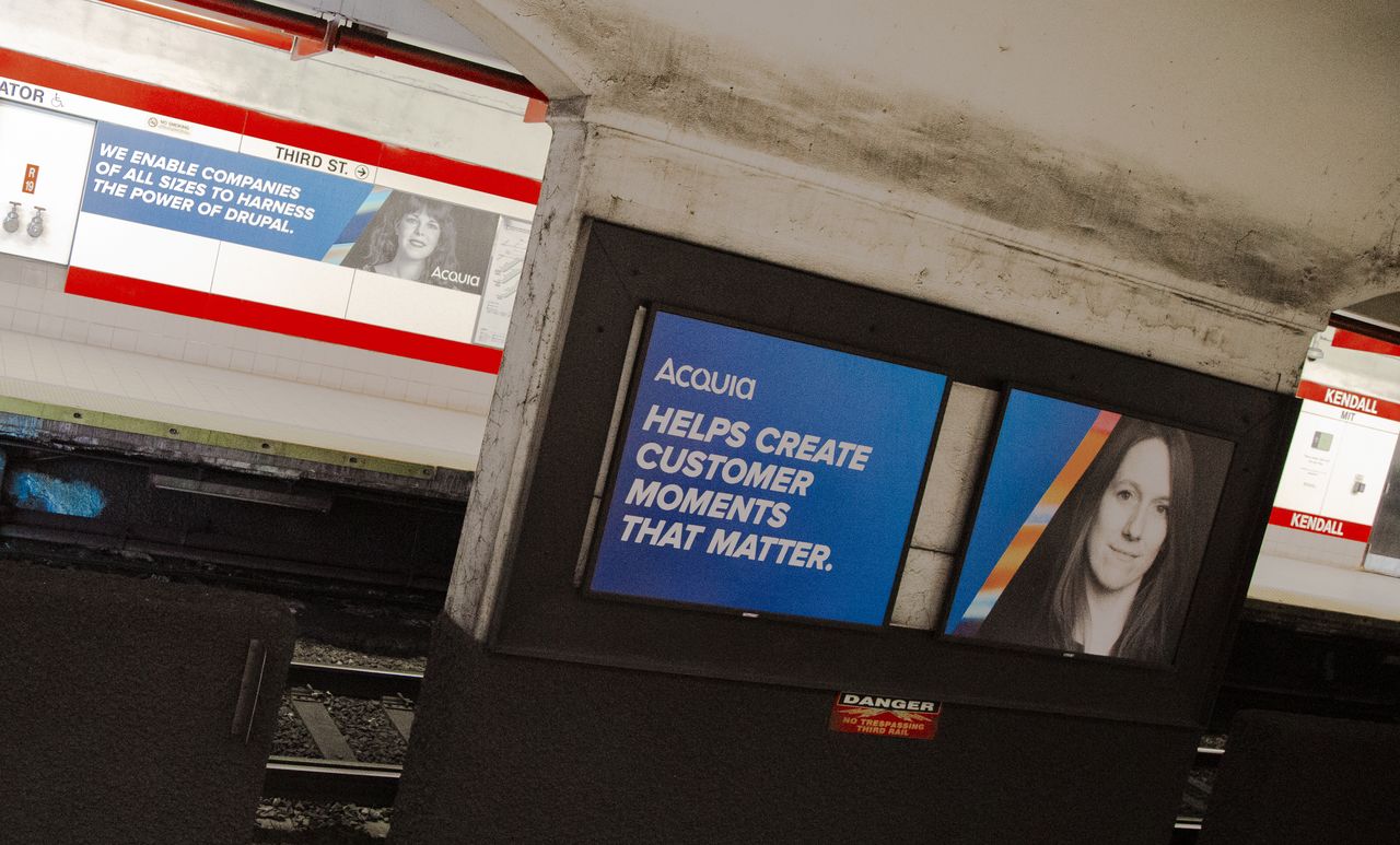 Acquia advertisements with portraits and slogans are displayed at Kendall Square subway station, promoting customer engagement and Drupal technology.
