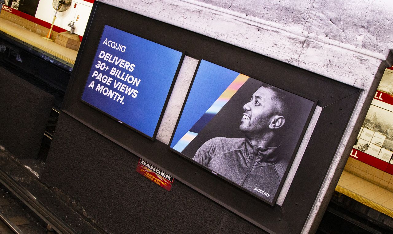 Two Acquia advertisements at a subway station, one highlighting page views and the other showing a smiling man.