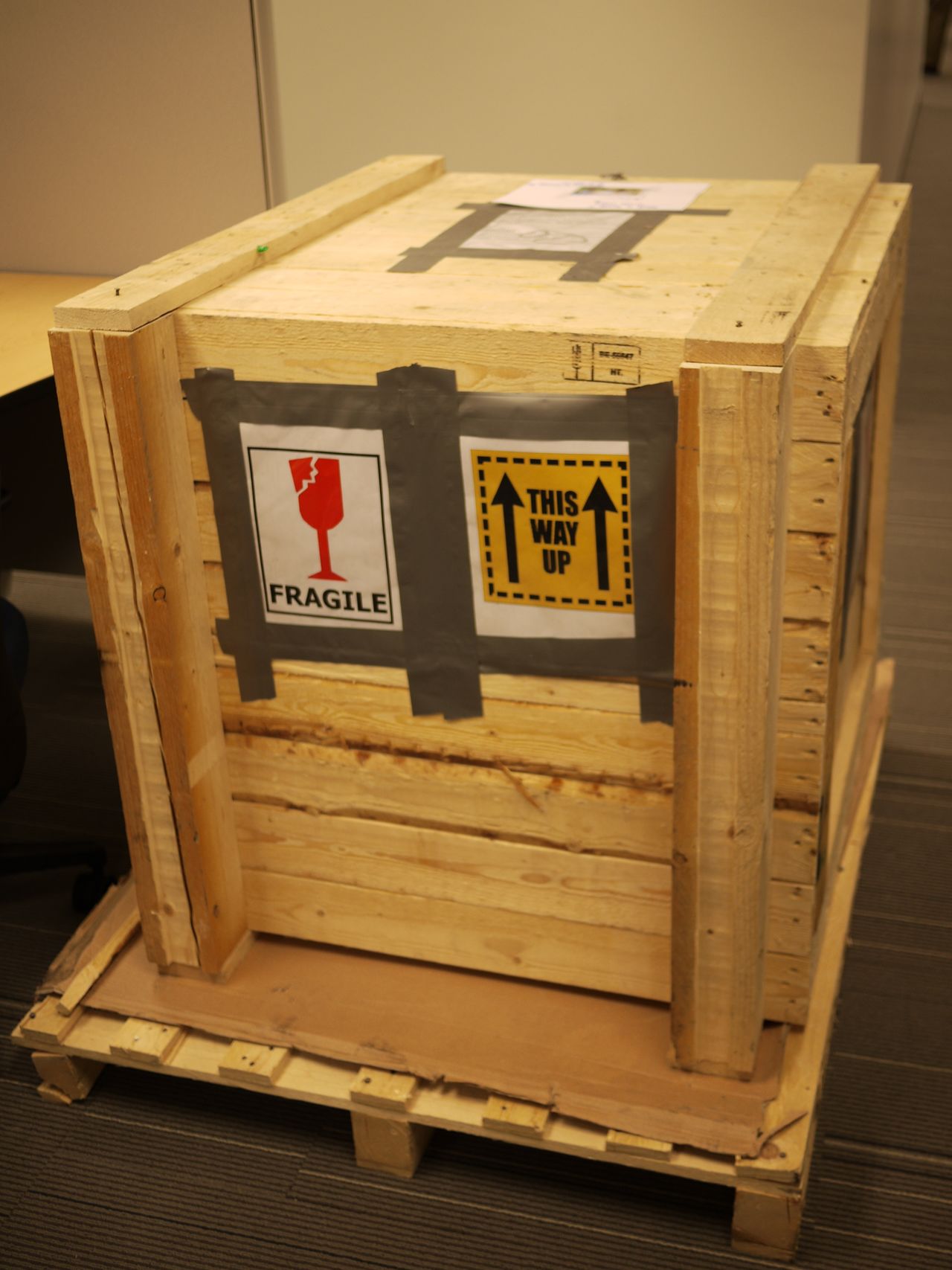 A large wooden crate with "Fragile" and "This Way Up" signs taped to its side, placed indoors on a pallet.