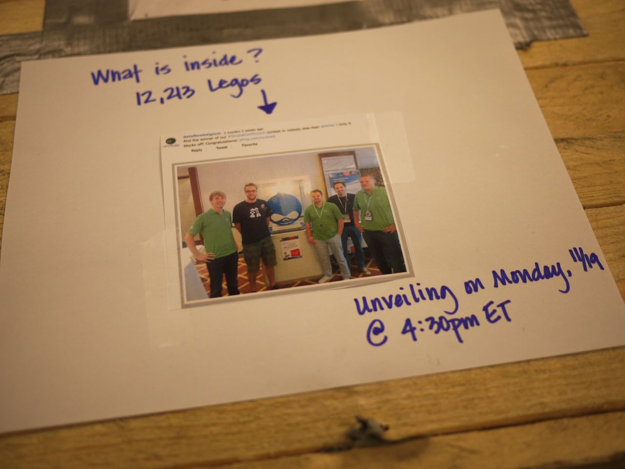 A printed photo shows five people standing next to a Lego Druplicon sculpture, with handwritten event details around it.