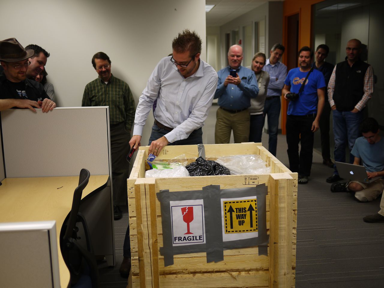 A man opens a large wooden crate labeled "Fragile" while a group of people watches in an office.