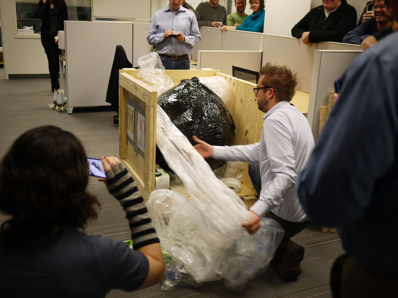 A man unwraps a large, black plastic-covered object from a wooden crate while others watch and take photos.