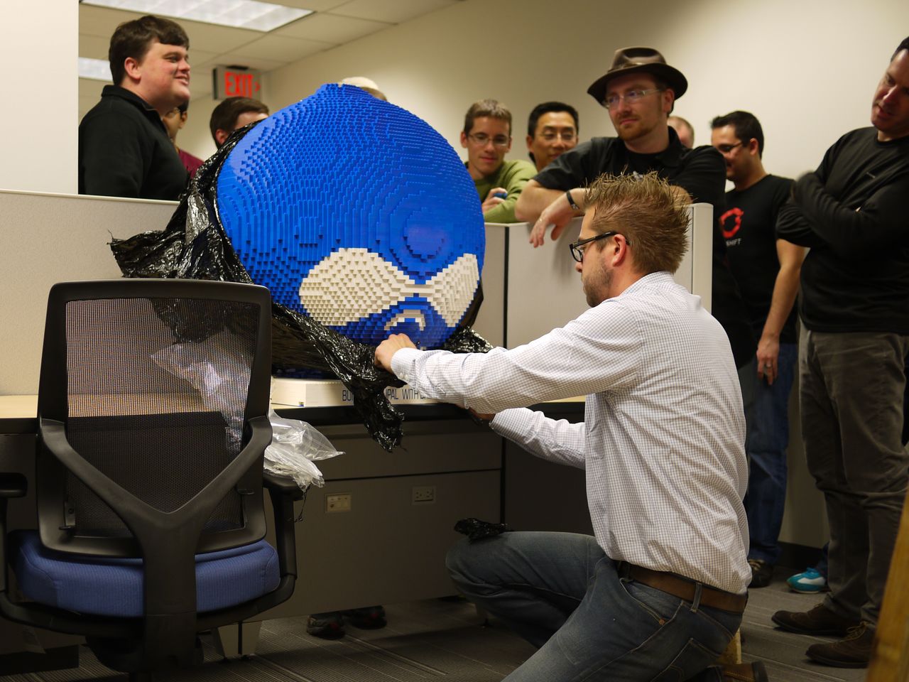 A man unwraps a large blue Lego Druplicon sculpture while a group of people watches in an office.