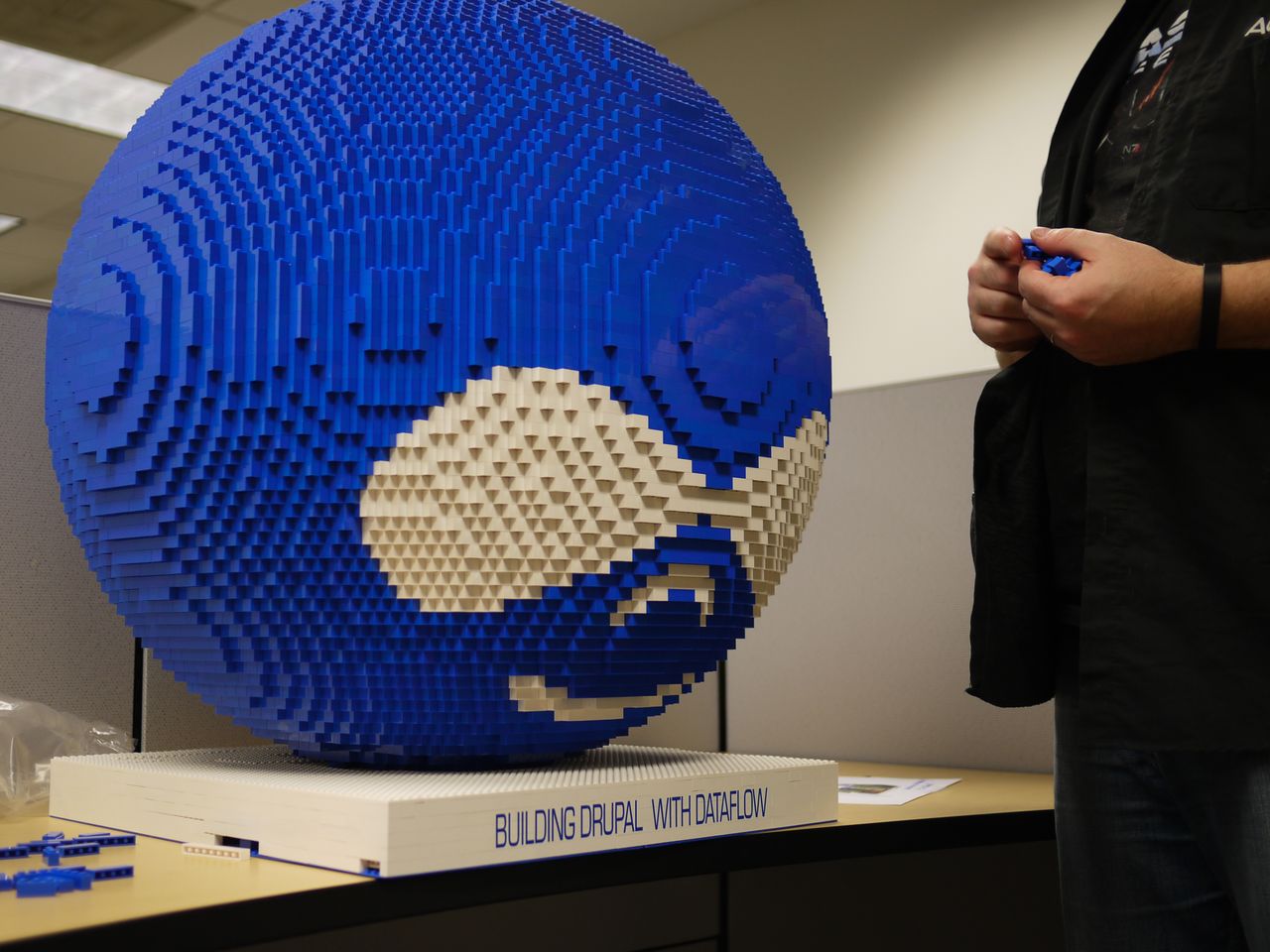 A large Lego sculpture of the Drupal logo sits on a table while a person adds blue bricks.