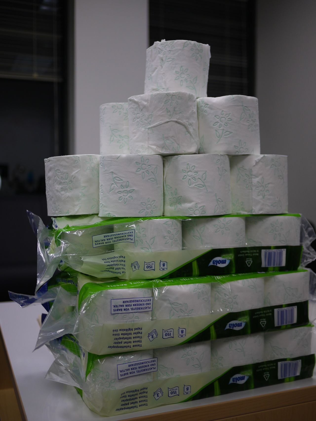 A pyramid-shaped stack of toilet paper rolls is arranged on a table, with packaged rolls at the base.