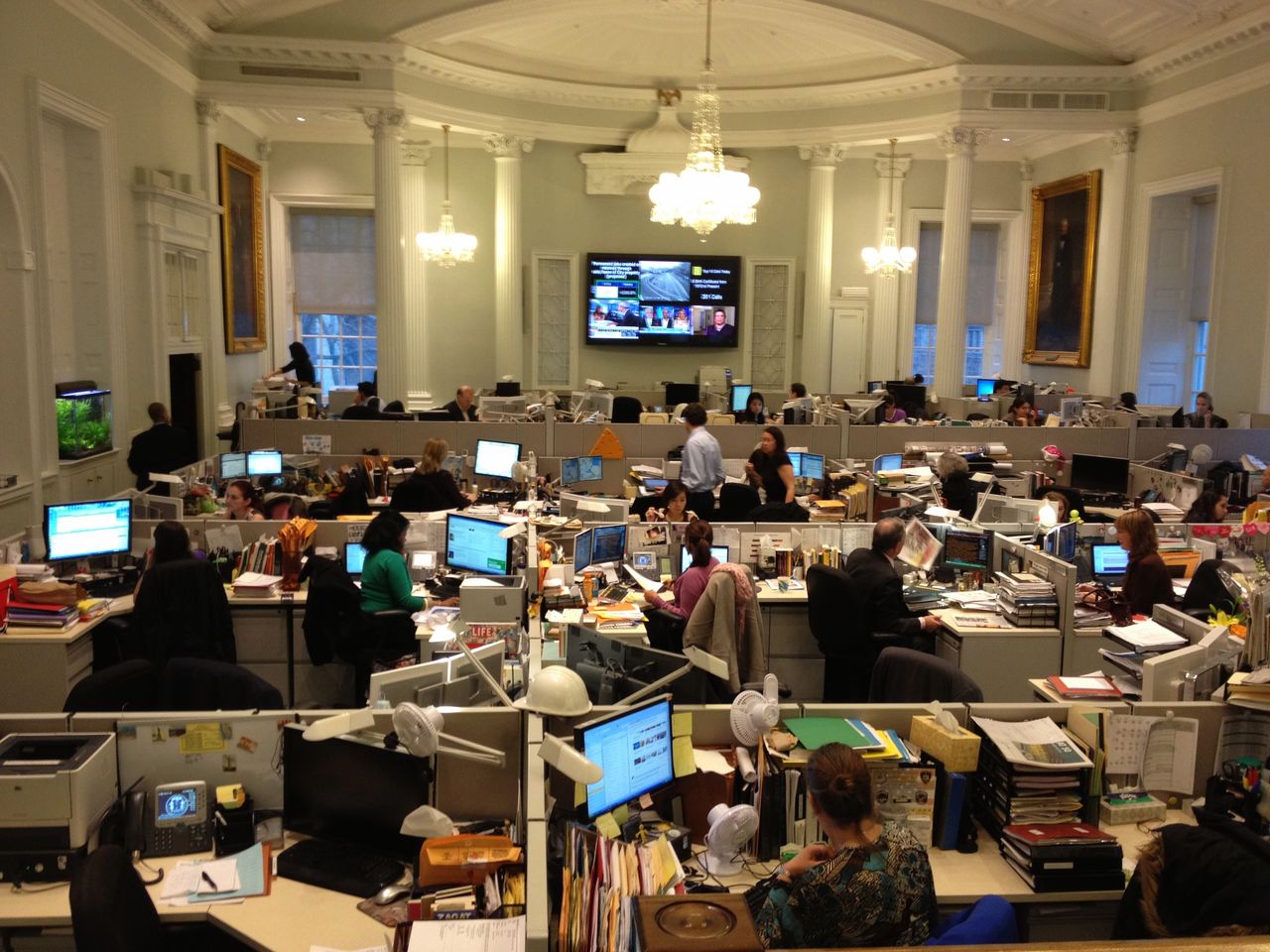 An office with employees working at desks, using computers, and having discussions in a well-lit, busy environment.
