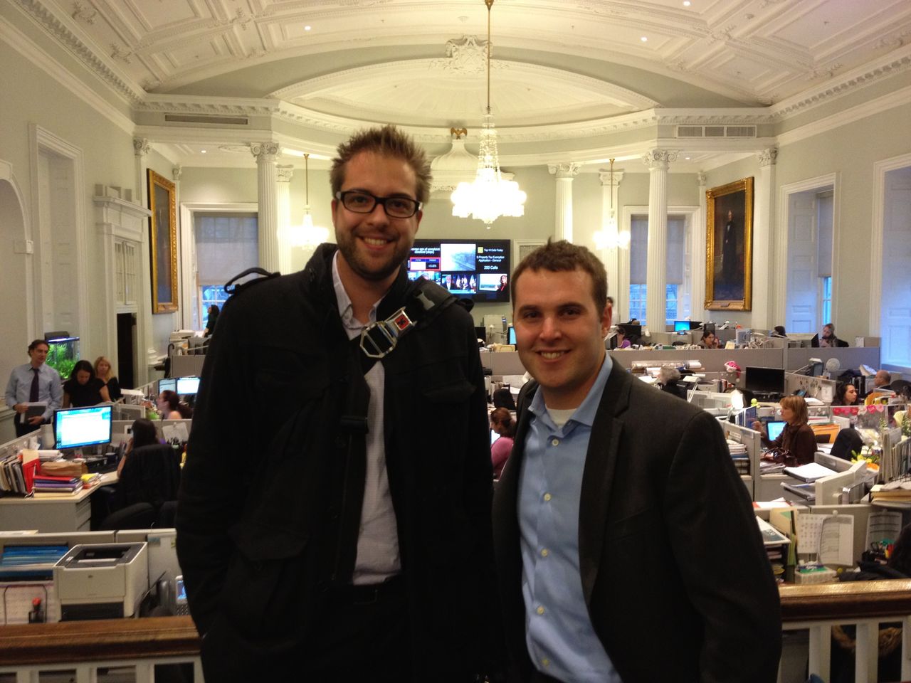 Two men smiling in an office with desks, computers, and people working in the background.