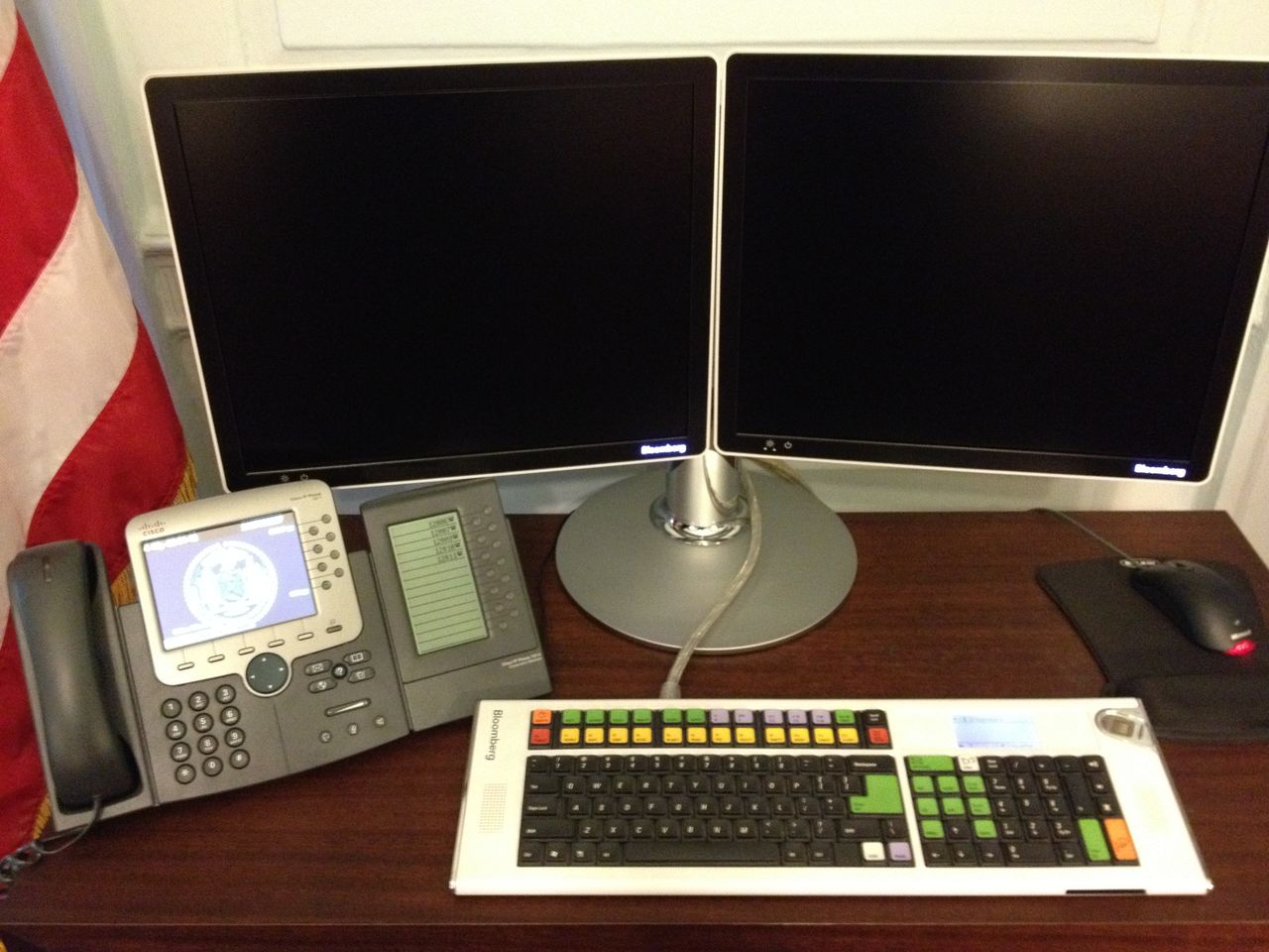 A desk with two blank monitors, a Bloomberg keyboard, a telephone, and a mouse on a pad.