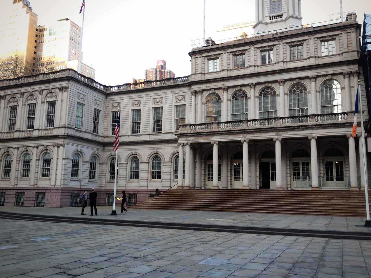 New York City Hall with American and city flags, and a few people walking on the paved plaza in front.