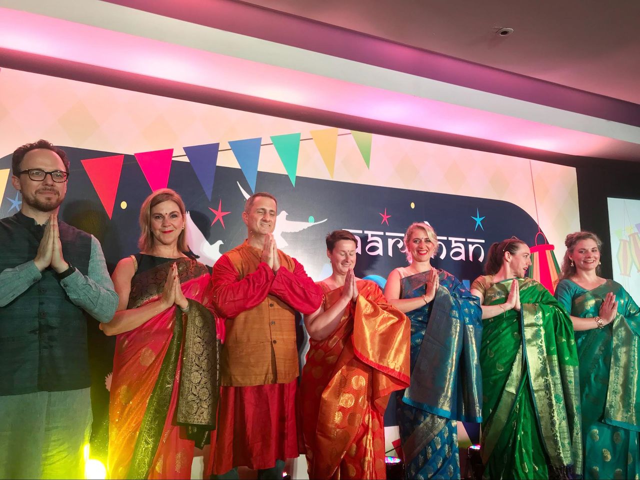 A group of people in traditional Indian attire stand on stage, smiling and greeting with folded hands.