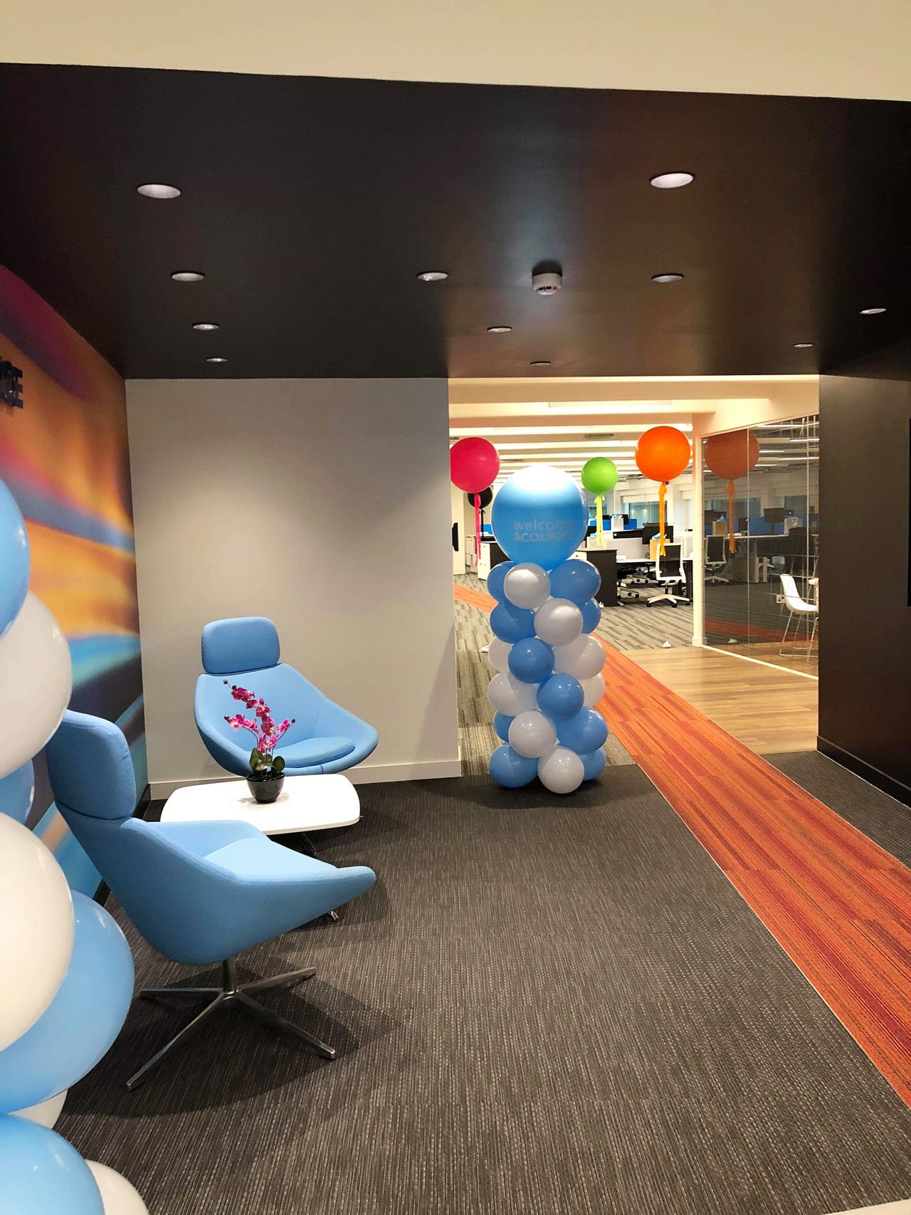A modern office lounge with blue chairs, a small table, and balloon decorations leading to an open workspace.