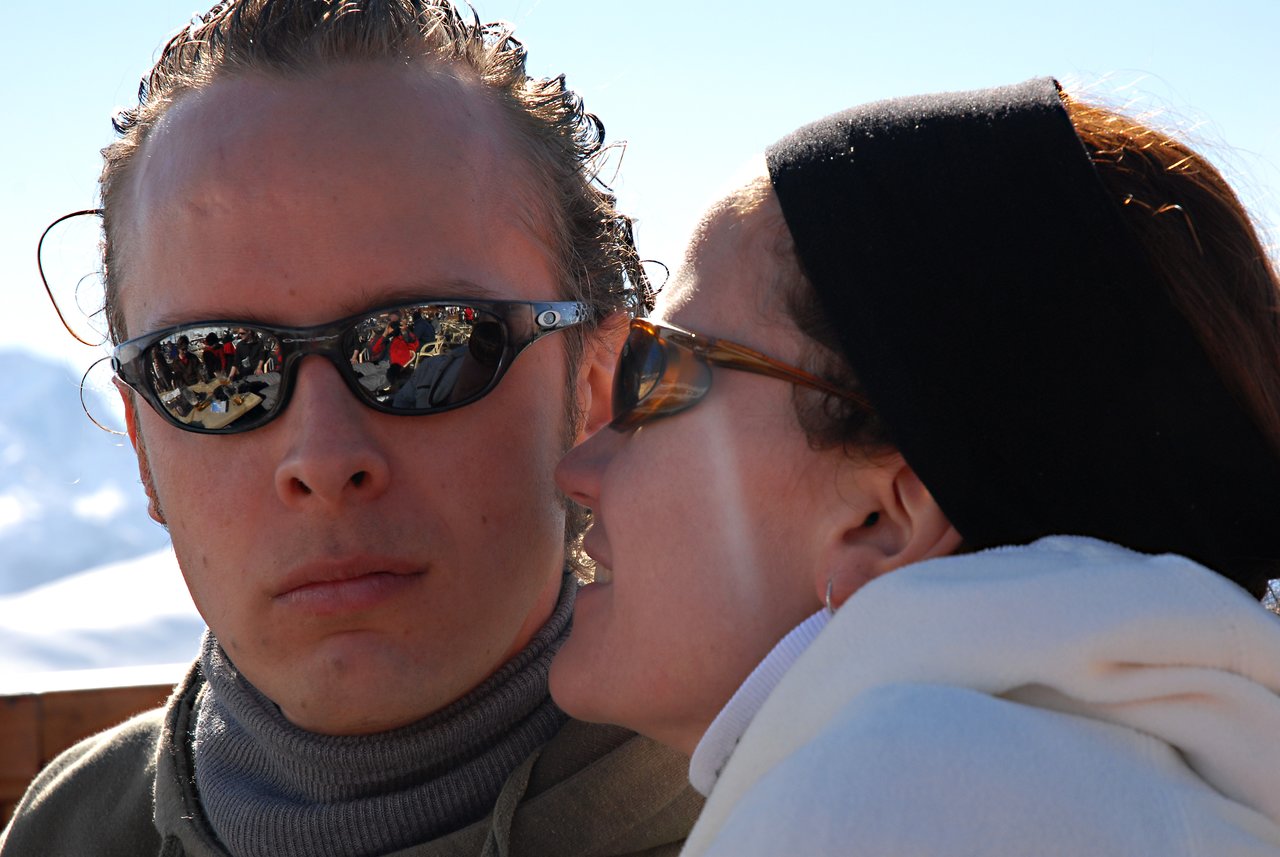 A woman leans in to speak closely to a man wearing sunglasses, who looks straight ahead with a neutral expression.