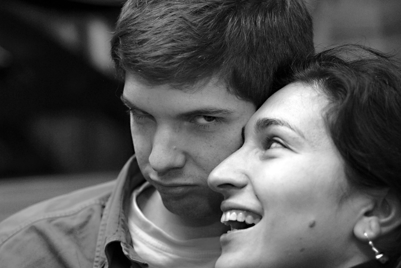 A man makes a playful pout while a woman leans in, smiling and laughing.