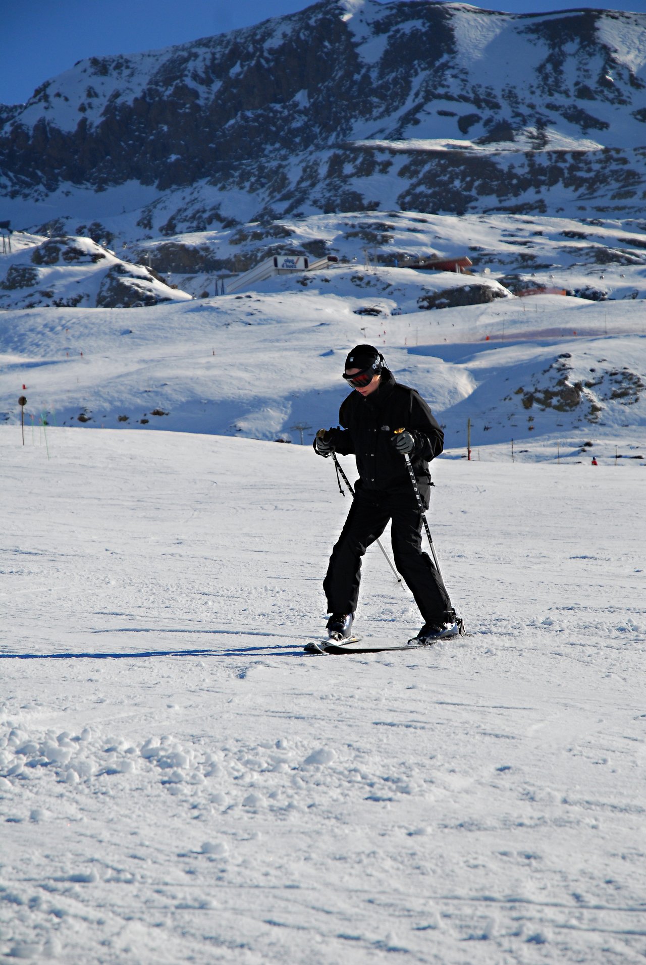 A person in black ski gear cautiously moves on skis down a snowy slope, holding ski poles for balance.