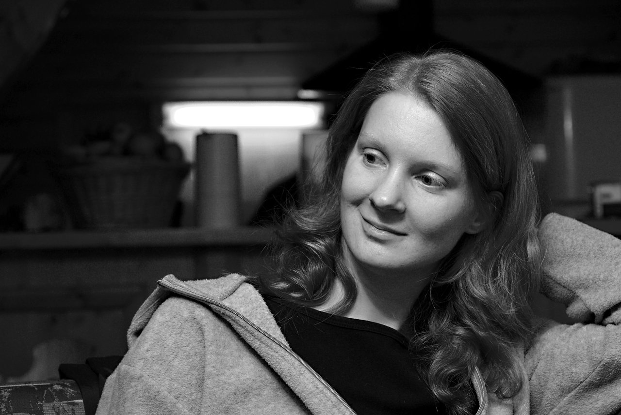 A woman with wavy hair sits indoors, wearing a zip-up jacket and smiling while looking to the side.